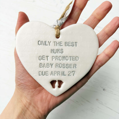 Pearlised white clay hanging heart with baby feet cut out of the bottom and grey personalisation, the heart is personalised with ONLY THE BEST MUMS GET PROMOTED BABY ROSSER DUE APRIL 27