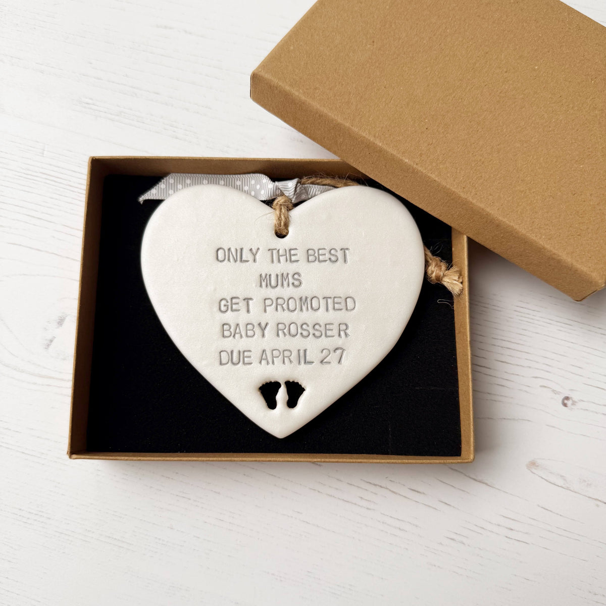 Pearlised white clay hanging heart with baby feet cut out of the bottom and grey personalisation, the heart is personalised with ONLY THE BEST MUMS GET PROMOTED BABY ROSSER DUE APRIL 27
