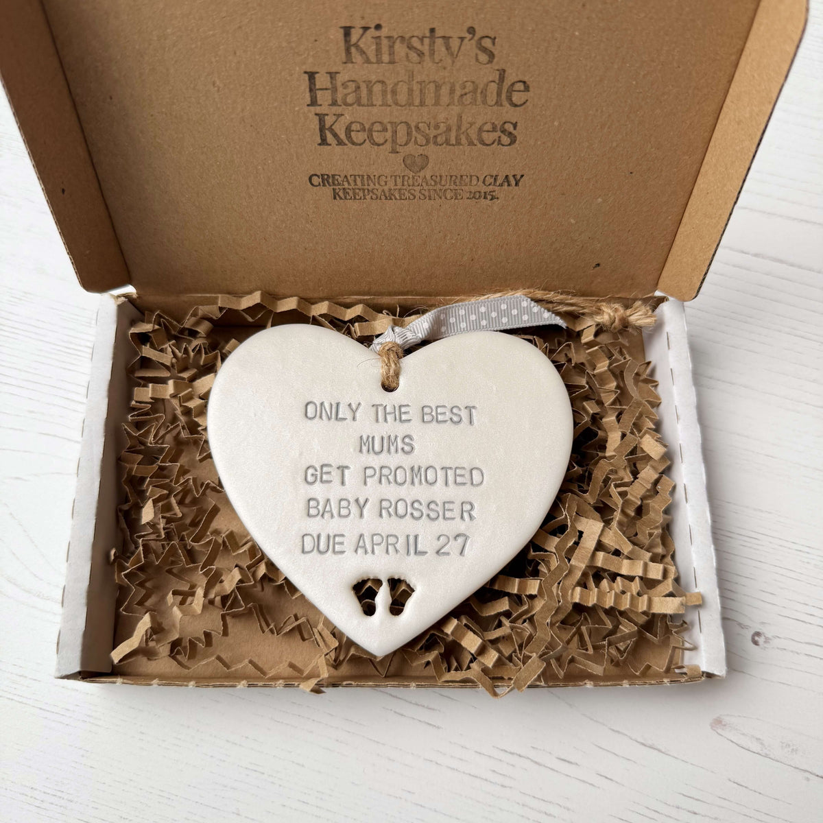 Pearlised white clay hanging heart with baby feet cut out of the bottom and grey personalisation, the heart is personalised with ONLY THE BEST MUMS GET PROMOTED BABY ROSSER DUE APRIL 27