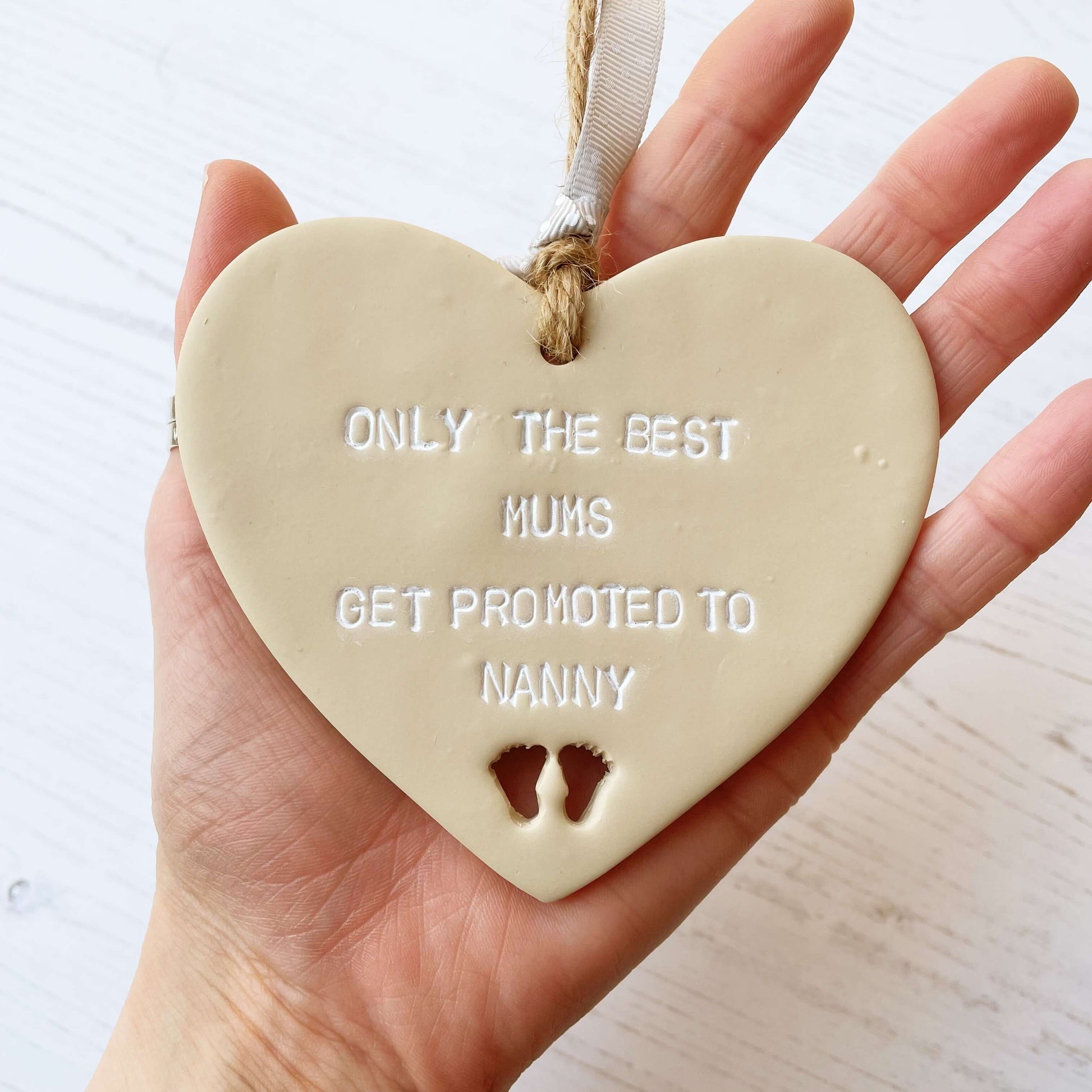 Beige clay hanging heart with baby feet cut out of the bottom and white personalisation, the heart is personalised with ONLY THE BEST MUMS GET PROMOTED TO NANNY