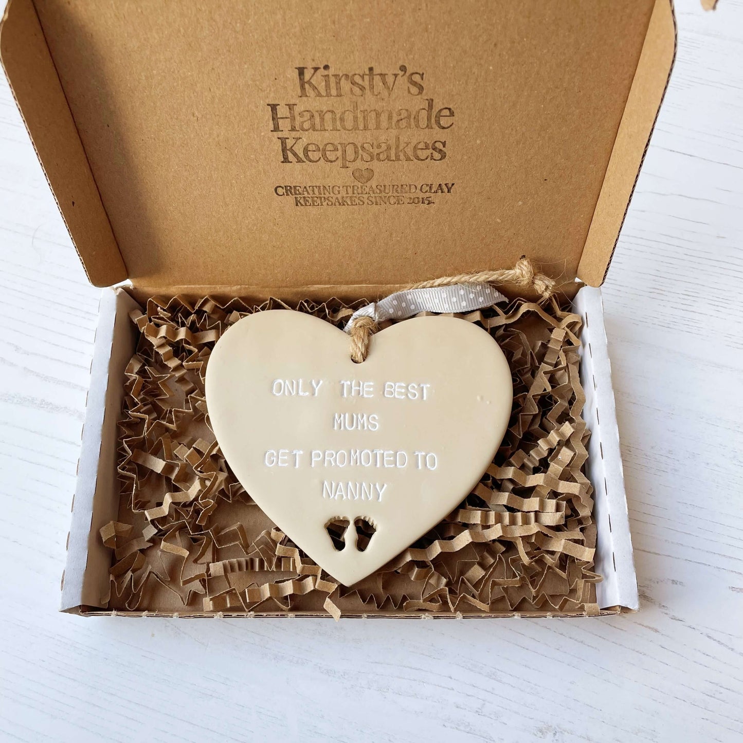 Beige clay hanging heart with baby feet cut out of the bottom and white personalisation, the heart is personalised with ONLY THE BEST MUMS GET PROMOTED TO NANNY