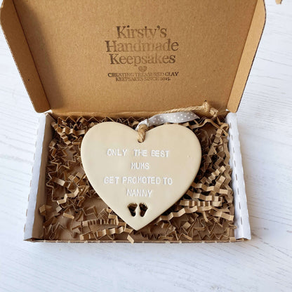 Beige clay hanging heart with baby feet cut out of the bottom and white personalisation, the heart is personalised with ONLY THE BEST MUMS GET PROMOTED TO NANNY