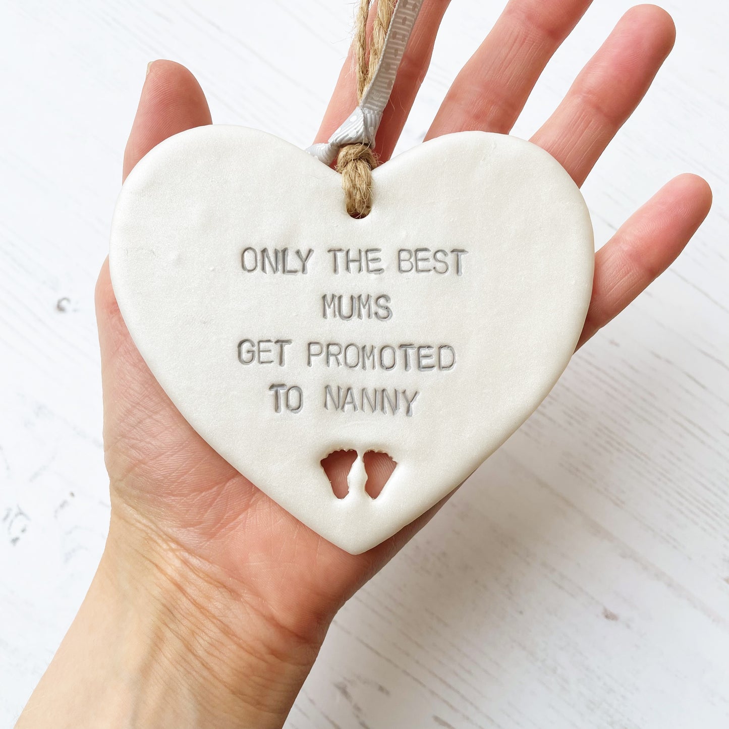 Pearlised white clay hanging heart with baby feet cut out of the bottom and grey personalisation, the heart is personalised with ONLY THE BEST MUMS GET PROMOTED TO NANNY