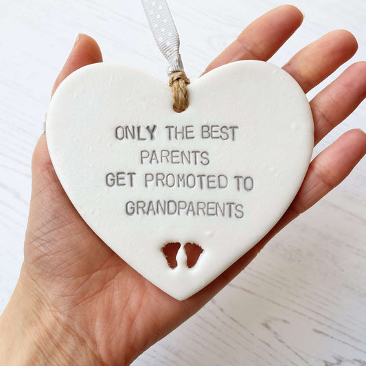 Pearlised white clay hanging heart with baby feet cut out of the bottom and grey personalisation, the heart is personalised with ONLY THE BEST PARENTS GET PROMOTED TO GRANDPARENTS