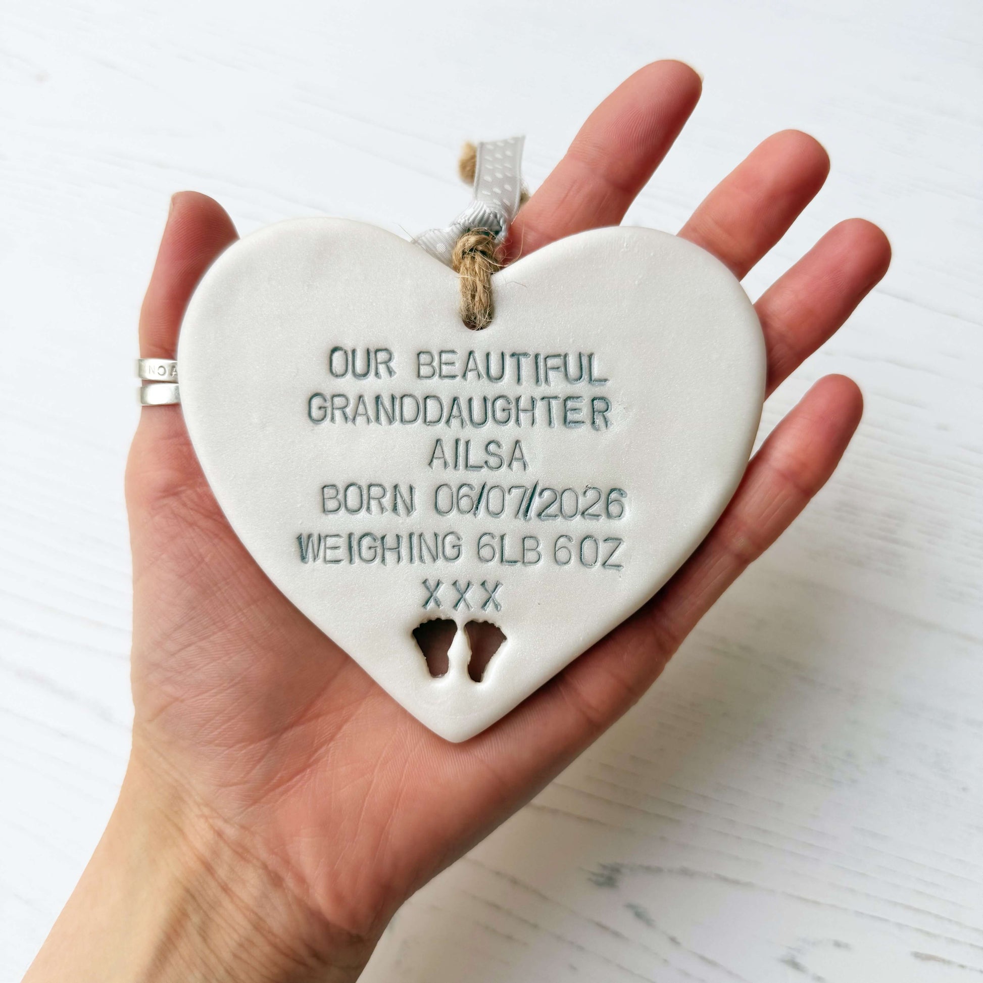 Pearlised white clay hanging heart with baby feet cut out of the bottom and grey personalisation, the heart is personalised with OUR BEAUTIFUL GRANDDAUGHTER AILSA BORN 06/07/2026 WEIGHING 6LB 6OZ XXX