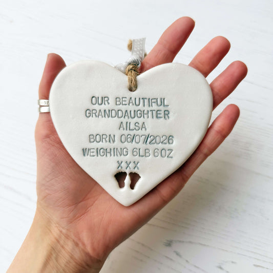 Pearlised white clay hanging heart with baby feet cut out of the bottom and grey personalisation, the heart is personalised with OUR BEAUTIFUL GRANDDAUGHTER AILSA BORN 06/07/2026 WEIGHING 6LB 6OZ XXX