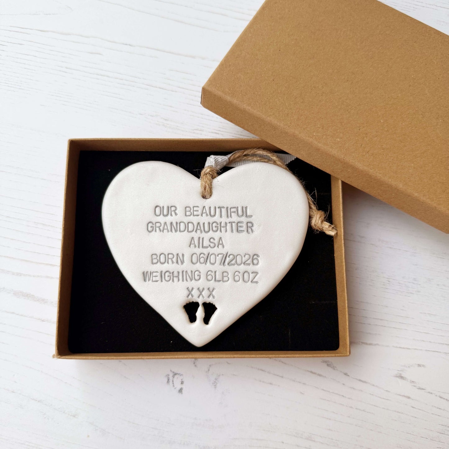 Pearlised white clay hanging heart with baby feet cut out of the bottom and grey personalisation, the heart is personalised with OUR BEAUTIFUL GRANDDAUGHTER AILSA BORN 06/07/2026 WEIGHING 6LB 6OZ XXX