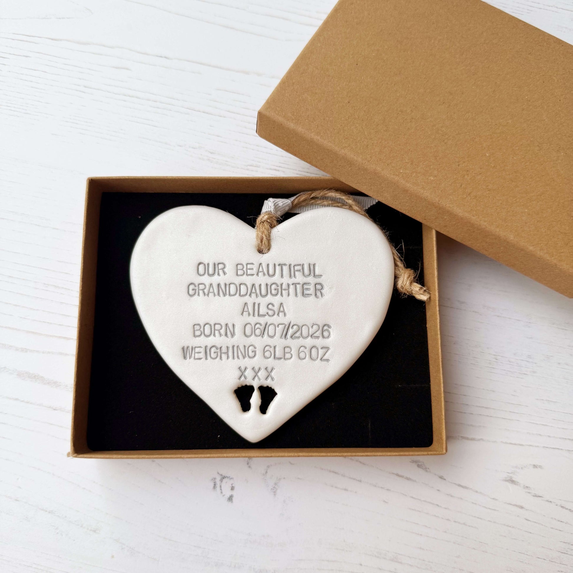Pearlised white clay hanging heart with baby feet cut out of the bottom and grey personalisation, the heart is personalised with OUR BEAUTIFUL GRANDDAUGHTER AILSA BORN 06/07/2026 WEIGHING 6LB 6OZ XXX