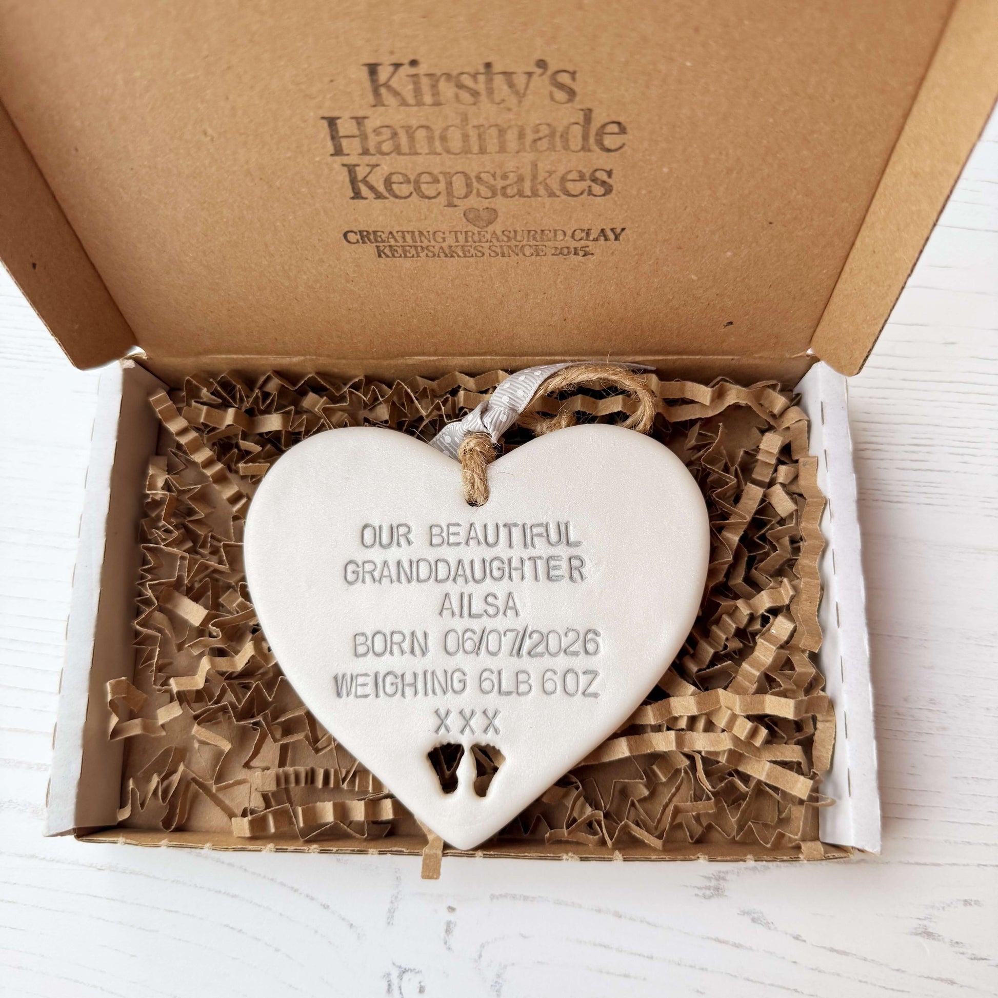 Pearlised white clay hanging heart with baby feet cut out of the bottom and grey personalisation, the heart is personalised with OUR BEAUTIFUL GRANDDAUGHTER AILSA BORN 06/07/2026 WEIGHING 6LB 6OZ XXX