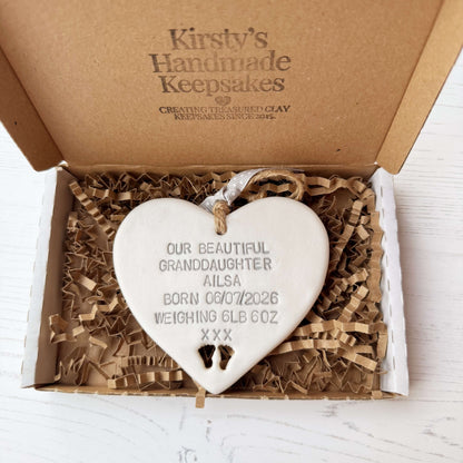 Pearlised white clay hanging heart with baby feet cut out of the bottom and grey personalisation, the heart is personalised with OUR BEAUTIFUL GRANDDAUGHTER AILSA BORN 06/07/2026 WEIGHING 6LB 6OZ XXX