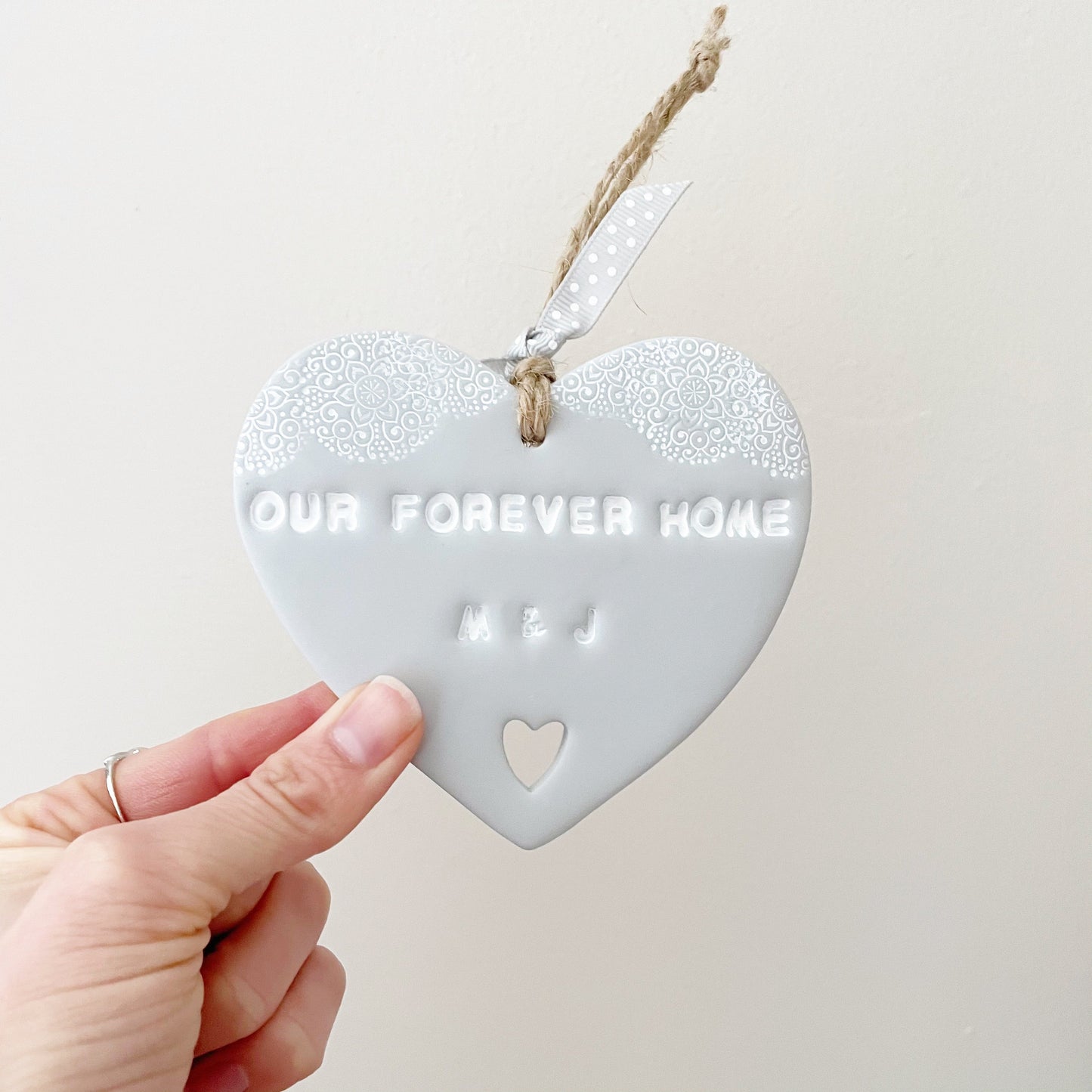 Personalised housewarming new home gift, grey clay heart with a white lace edge at the top of the heart and a heart cut out at the bottom with jute twine for hanging, the heart is personalised with OUR FOREVER HOME M & J