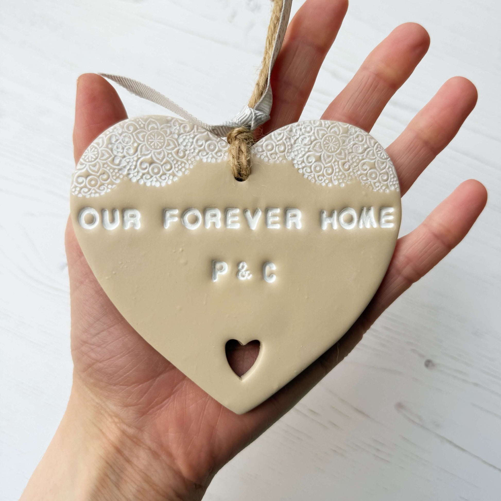 Personalised housewarming new home gift, beige clay heart with a white lace edge at the top of the heart and a heart cut out at the bottom with jute twine for hanging, the heart is personalised with OUR FOREVER HOME P & C