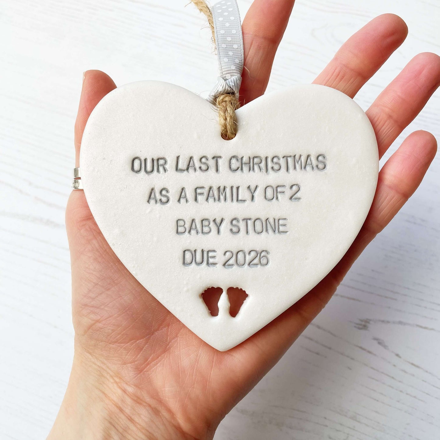 Pearlised white clay hanging heart pregnancy reveal with baby feet cut out of the bottom and grey personalisation, the heart is personalised with OUR LAST CHRISTMAS AS A FAMILY OF 2 BABY STONE DUE 2026