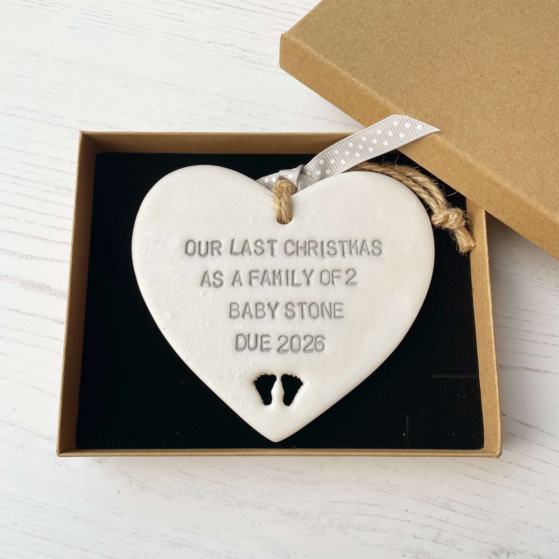 Pearlised white clay hanging heart pregnancy reveal with baby feet cut out of the bottom and grey personalisation, the heart is personalised with OUR LAST CHRISTMAS AS A FAMILY OF 2 BABY STONE DUE 2026