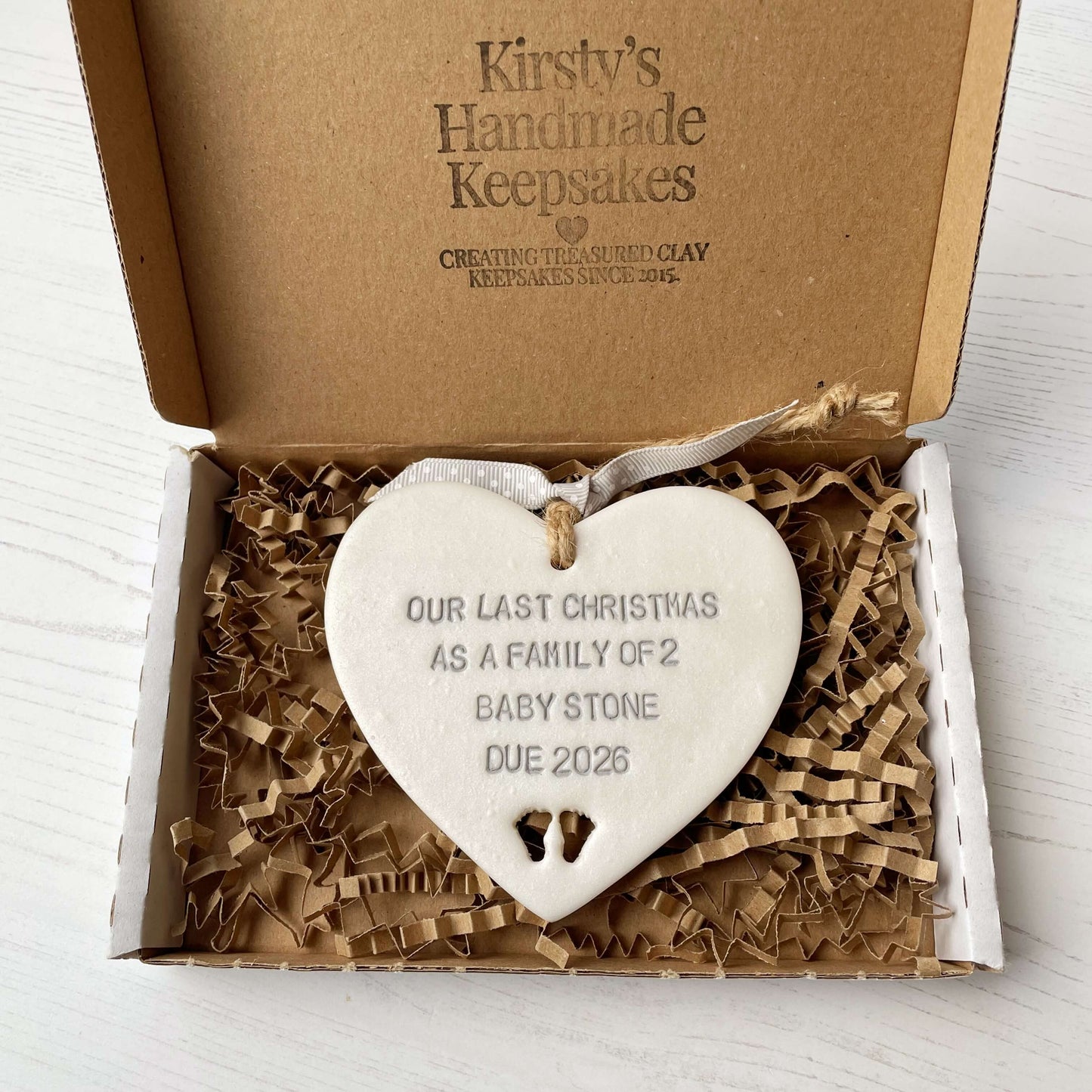 Pearlised white clay hanging heart pregnancy reveal with baby feet cut out of the bottom and grey personalisation, the heart is personalised with OUR LAST CHRISTMAS AS A FAMILY OF 2 BABY STONE DUE 2026