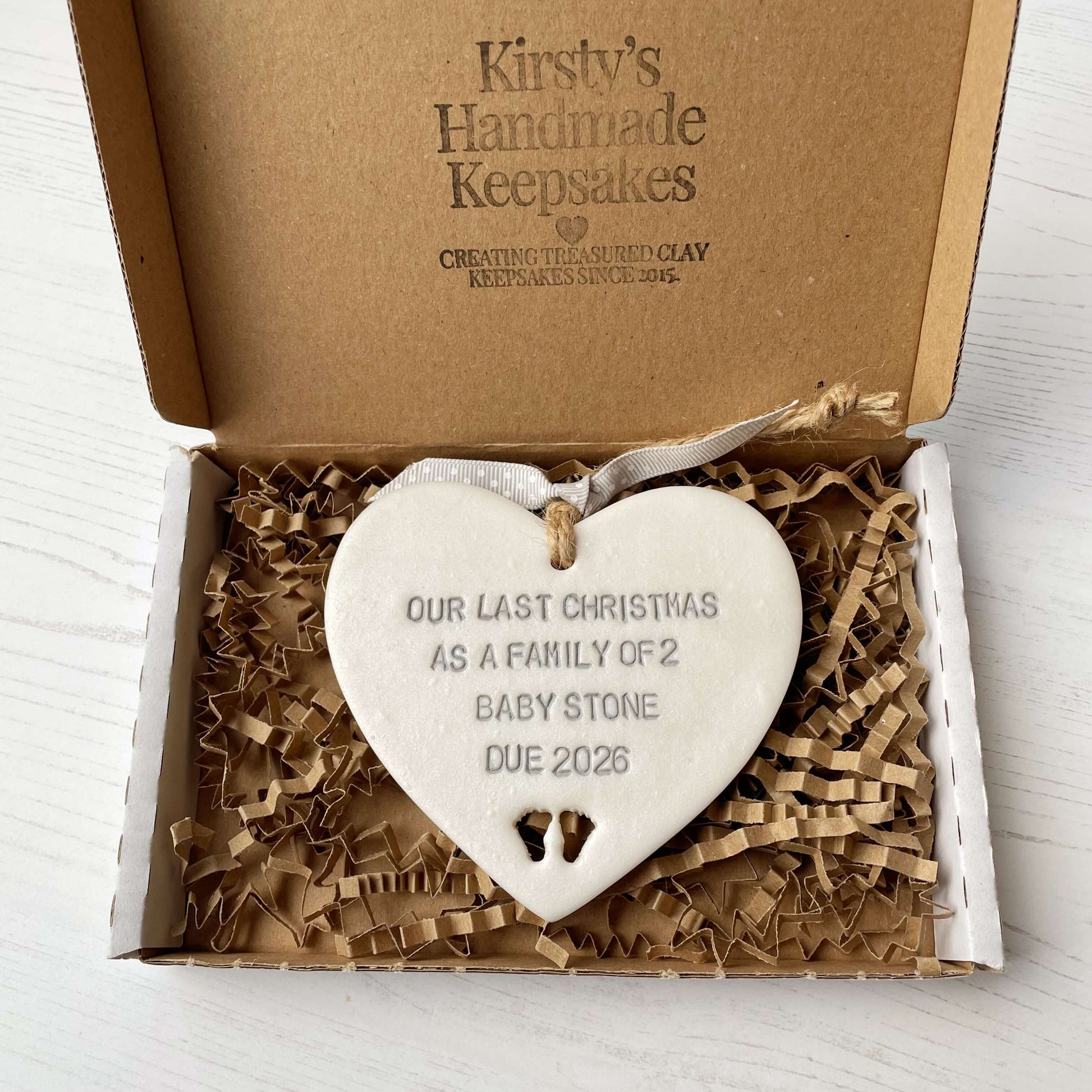Pearlised white clay hanging heart pregnancy reveal with baby feet cut out of the bottom and grey personalisation, the heart is personalised with OUR LAST CHRISTMAS AS A FAMILY OF 2 BABY STONE DUE 2026