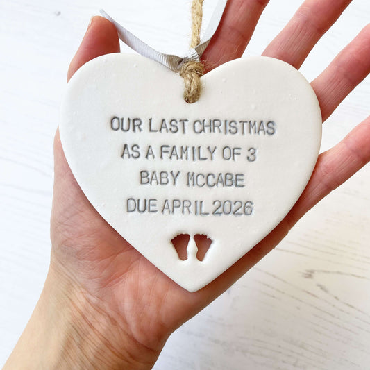 Pearlised white clay hanging heart pregnancy reveal with baby feet cut out of the bottom and grey personalisation, the heart is personalised with OUR LAST CHRISTMAS AS A FAMILY OF 3 BABY MACCABE DUE APRIL 2026