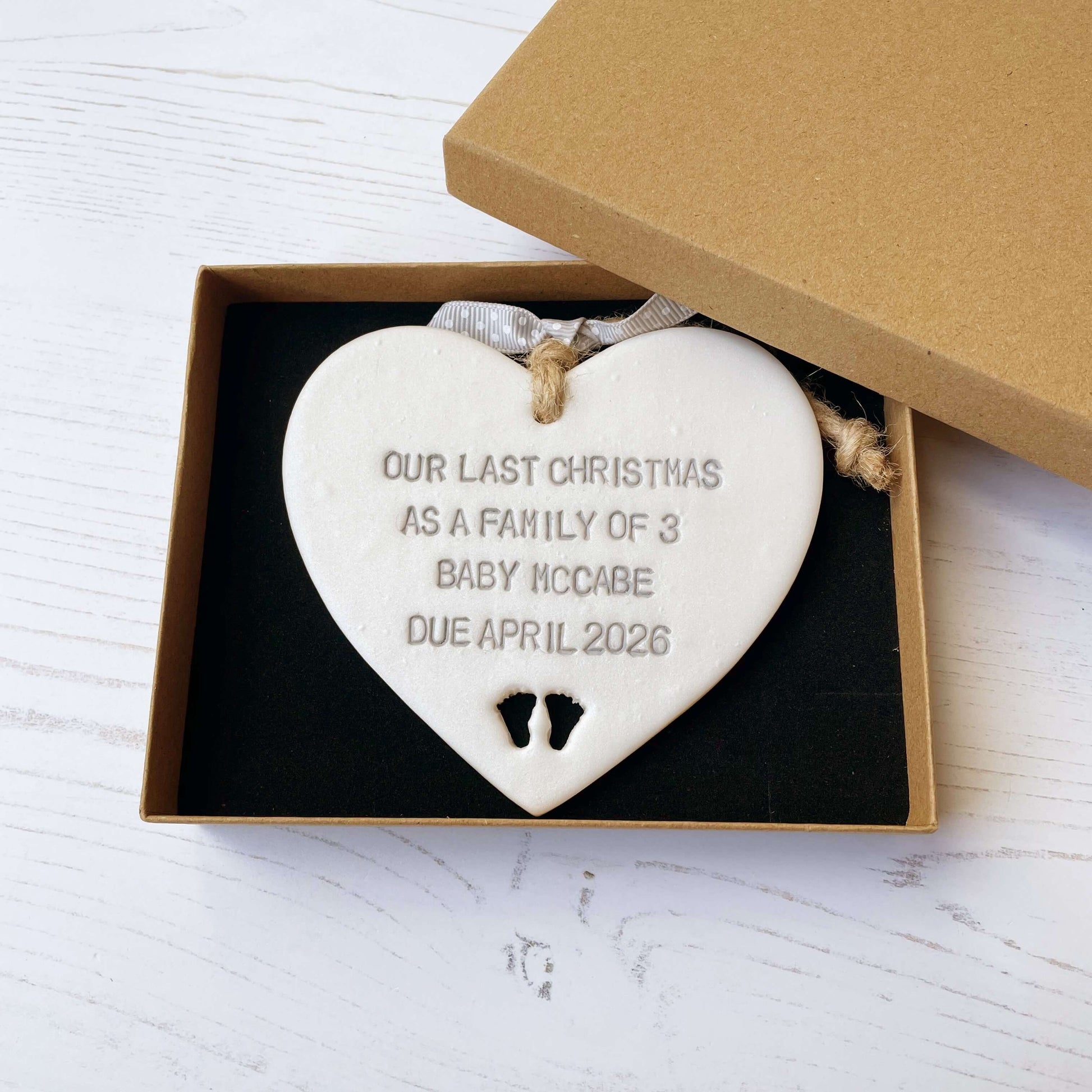 Pearlised white clay hanging heart pregnancy reveal with baby feet cut out of the bottom and grey personalisation, the heart is personalised with OUR LAST CHRISTMAS AS A FAMILY OF 3 BABY MACCABE DUE APRIL 2026