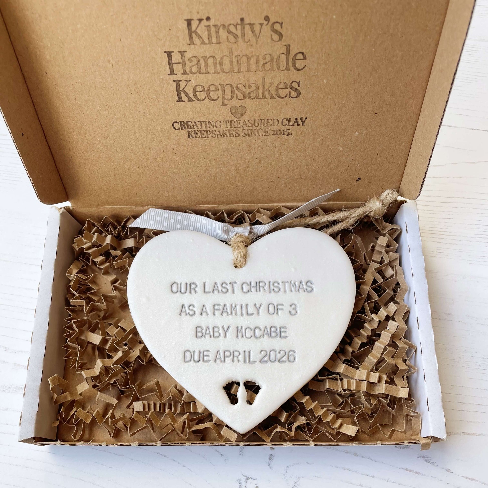 Pearlised white clay hanging heart pregnancy reveal with baby feet cut out of the bottom and grey personalisation, the heart is personalised with OUR LAST CHRISTMAS AS A FAMILY OF 3 BABY MACCABE DUE APRIL 2026