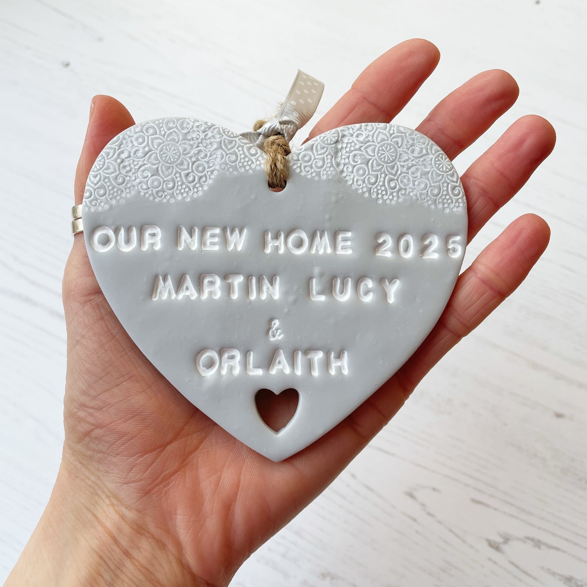 Personalised new home gift grey clay heart with a white lace edge at the top & a heart cut out at the bottom with twine for hanging personalised with OUR NEW HOME 2025 MARTIN LUCY &  ORLAITH