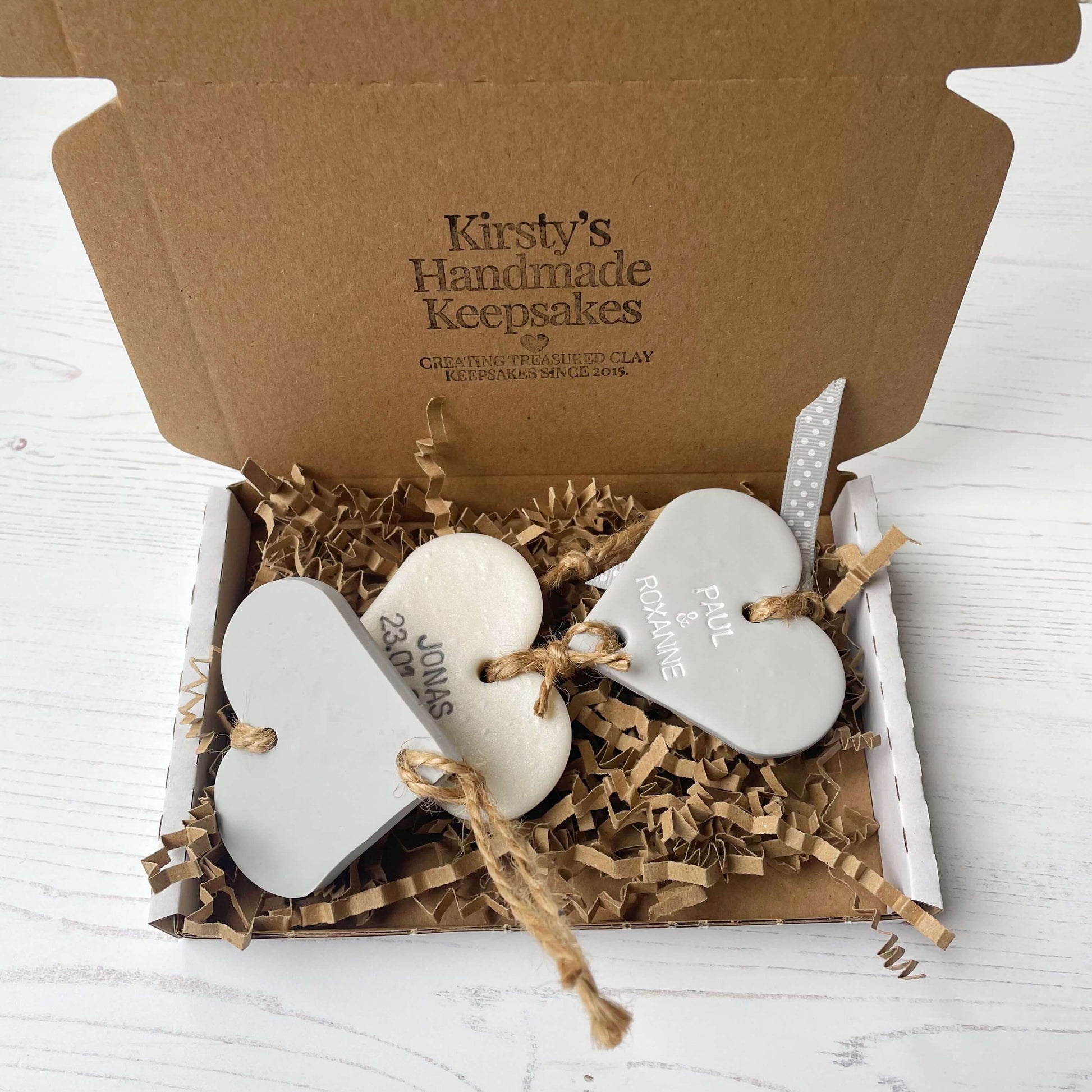 Family tree keepsake, grey small polymer clay heart with a pearlised white small heart hanging below, a small grey heart hanging at the bottom, personalised with family names and dates In a white postal box with shredded brown zigzag Kraft paper