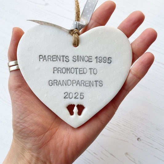 Pearlised white clay hanging heart with baby feet cut out of the bottom and grey personalisation, the heart is personalised with PARENTS SINCE 1995 PROMOTED TO GRANDPARENTS 2025