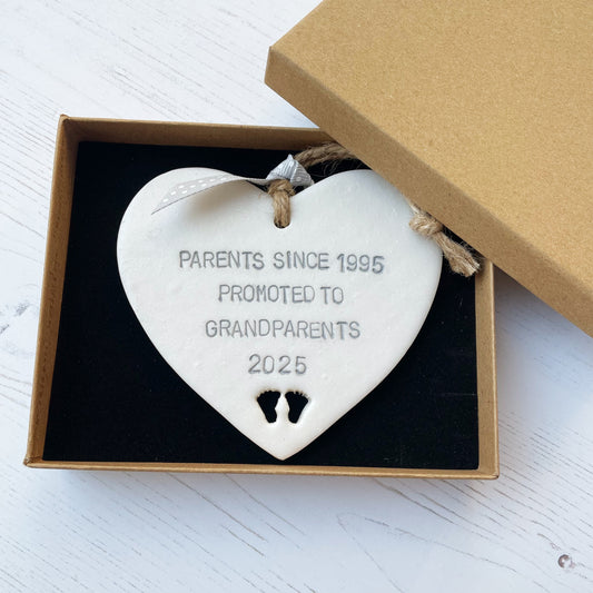 Pearlised white clay hanging heart with baby feet cut out of the bottom and grey personalisation, the heart is personalised with PARENTS SINCE 1995 PROMOTED TO GRANDPARENTS 2025