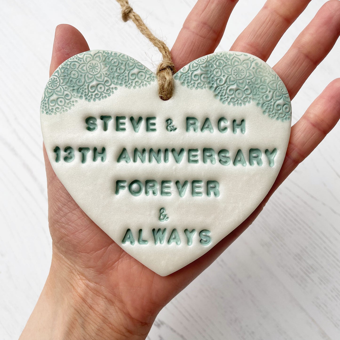 Personalised 13th wedding anniversary gift, pearlised white clay hanging heart with a sage green lace edge at the top of the heart, the heart is personalised with STEVE & RACH 13TH ANNIVERSARY FOREVER & ALWAYS
