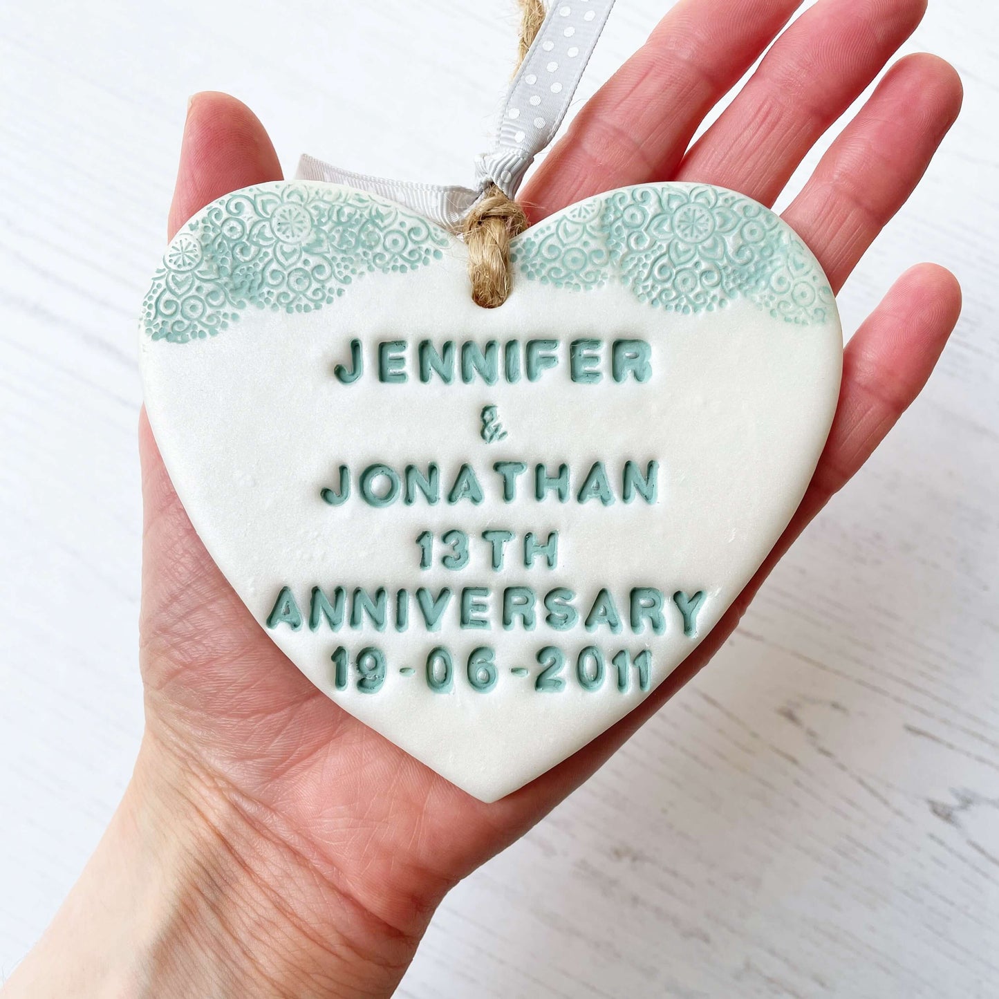 Personalised 13th anniversary gift, pearlised white clay hanging heart with a sage green lace edge at the top of the heart, the heart is personalised with JENNIFER & JONATHAN 13TH ANNIVERSARY 19-06-2011