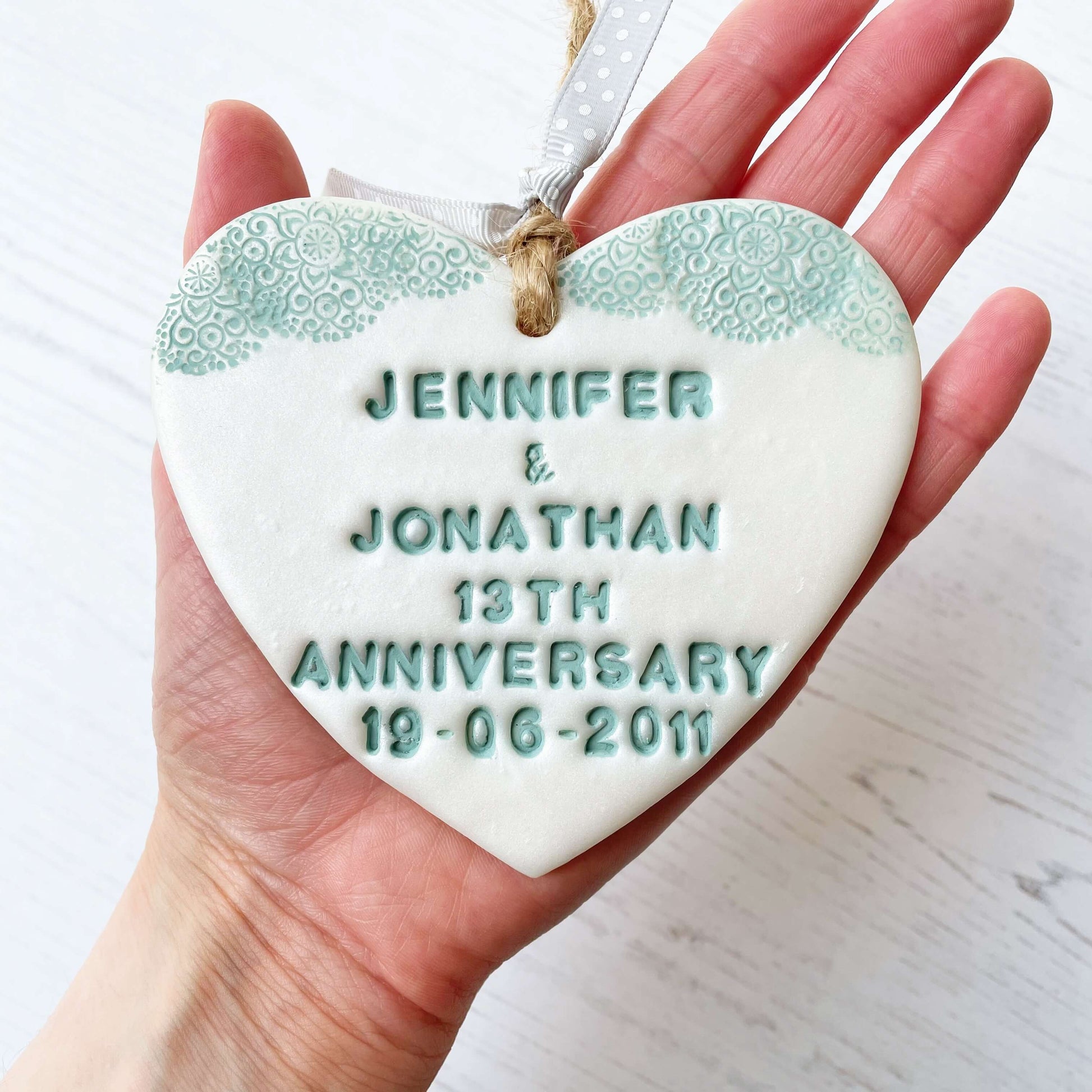 Personalised 13th anniversary gift, pearlised white clay hanging heart with a sage green lace edge at the top of the heart, the heart is personalised with JENNIFER & JONATHAN 13TH ANNIVERSARY 19-06-2011