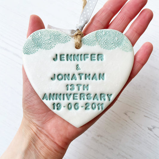 Personalised 13th anniversary gift, pearlised white clay hanging heart with a sage green lace edge at the top of the heart, the heart is personalised with JENNIFER & JONATHAN 13TH ANNIVERSARY 19-06-2011