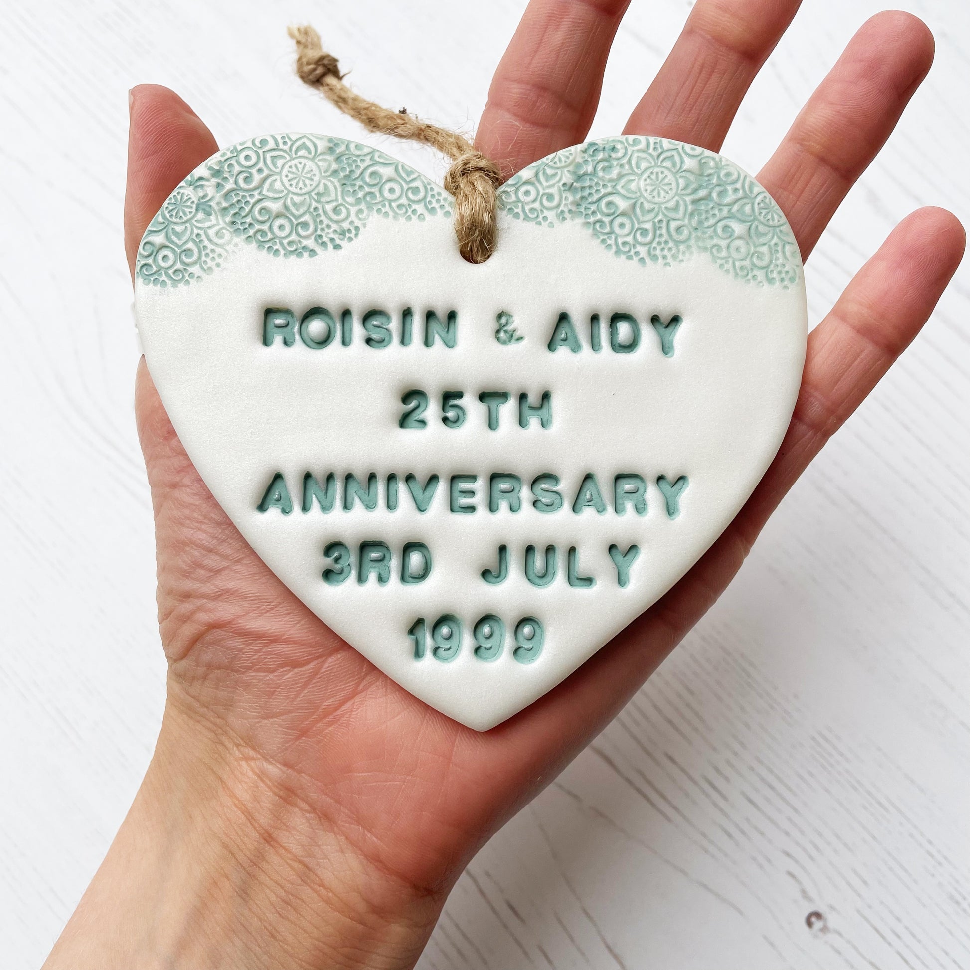 Personalised 25th anniversary gift, pearlised white clay hanging heart with a sage green lace edge at the top of the heart, the heart is personalised with ROISIN & AIDY 25TH ANNIVERSARY 3RD JULY 1999