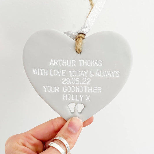 Personalised Christening gift, grey clay hanging heart with baby feet cut out of the bottom, the heart is personalised with  ARTHUR THOMAS WITH LOVE TODAY & ALWAYS 29.05.22 YOUR GODMOTHER HOLLY X