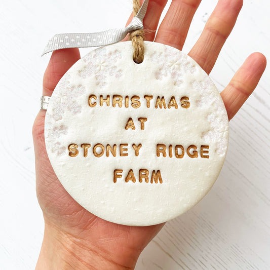 Personalised first Christmas in our new home bauble ornament, pearlised white round clay with CHRISTMAS AT STONEY RIDGE FARM in gold paint, decorated with 3 iridescent glitter snowflakes on either side of the bauble