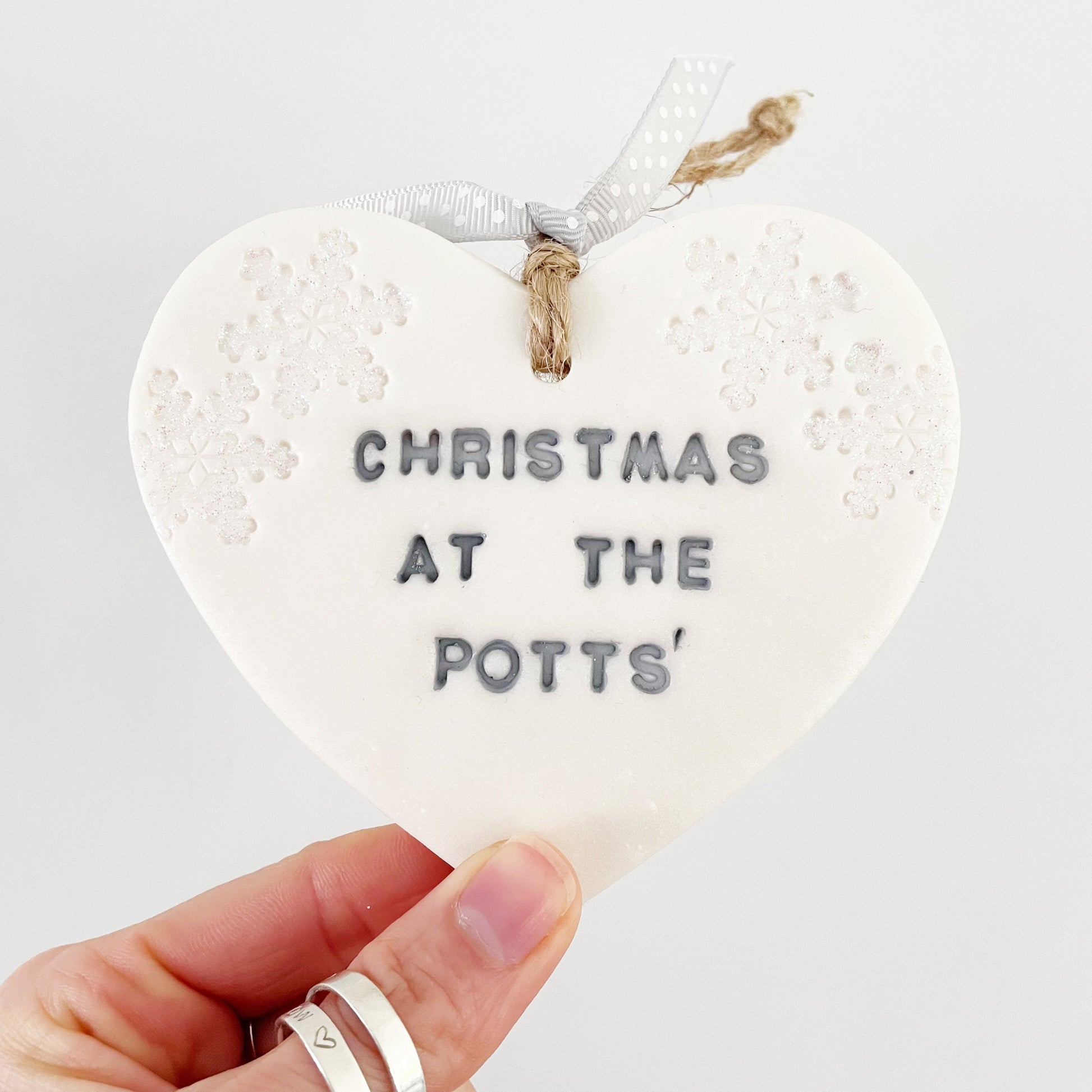 Personalised Christmas at the heart ornament, pearlised white clay with CHRISTMAS AT THE POTTS’ painted grey, decorated with 2 iridescent glitter snowflakes on either side of the top of the heart