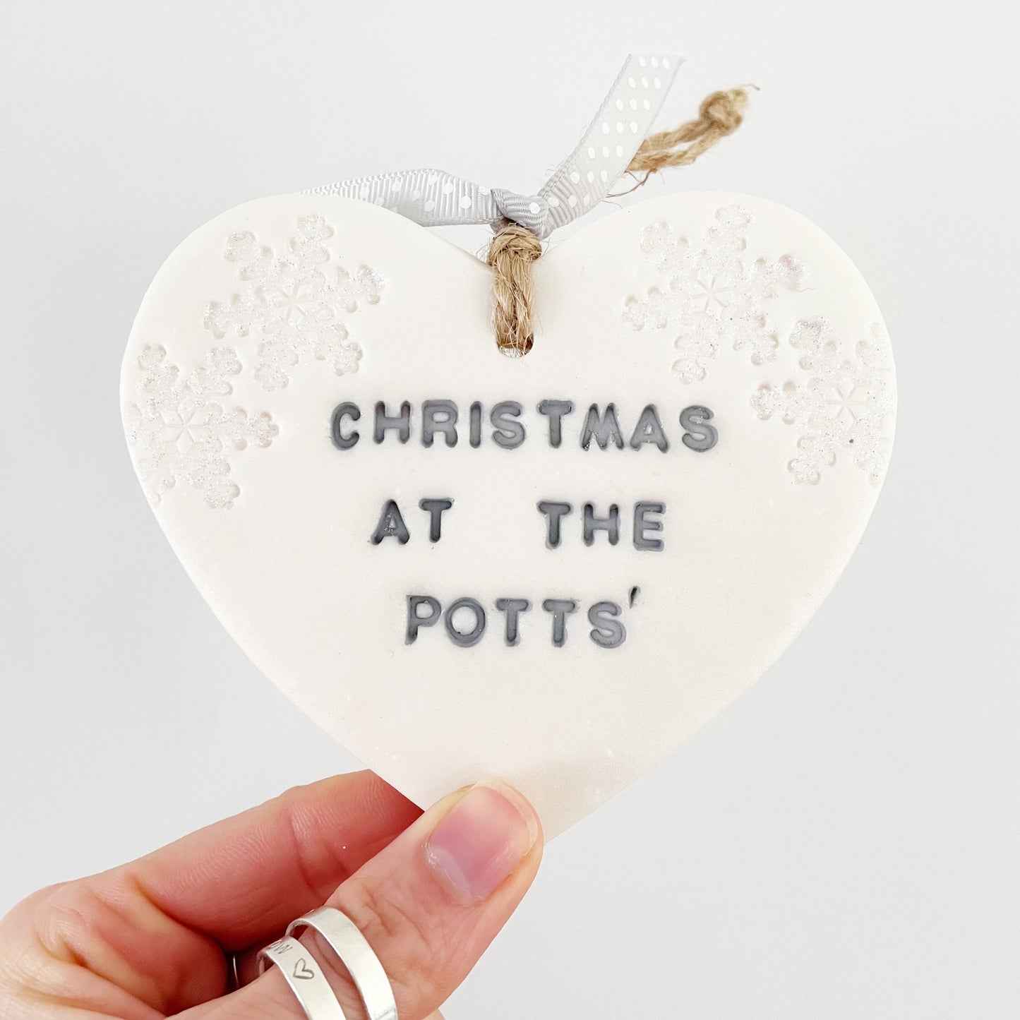 Personalised baby's first Christmas heart ornament, pearlised white clay with CHRISTMAS AT THE POTTS’ painted grey, decorated with 2 iridescent glitter snowflakes on either side of the top of the heart