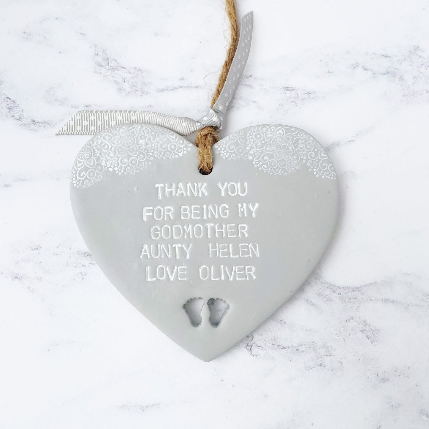 Personalised Godmother gift, grey clay hanging heart with baby feet cut out of the bottom and white lace edge at the top of the heart, the heart is personalised with THANK YOU FOR BEING MY GODMOTHER HELEN LOVE OLIVER