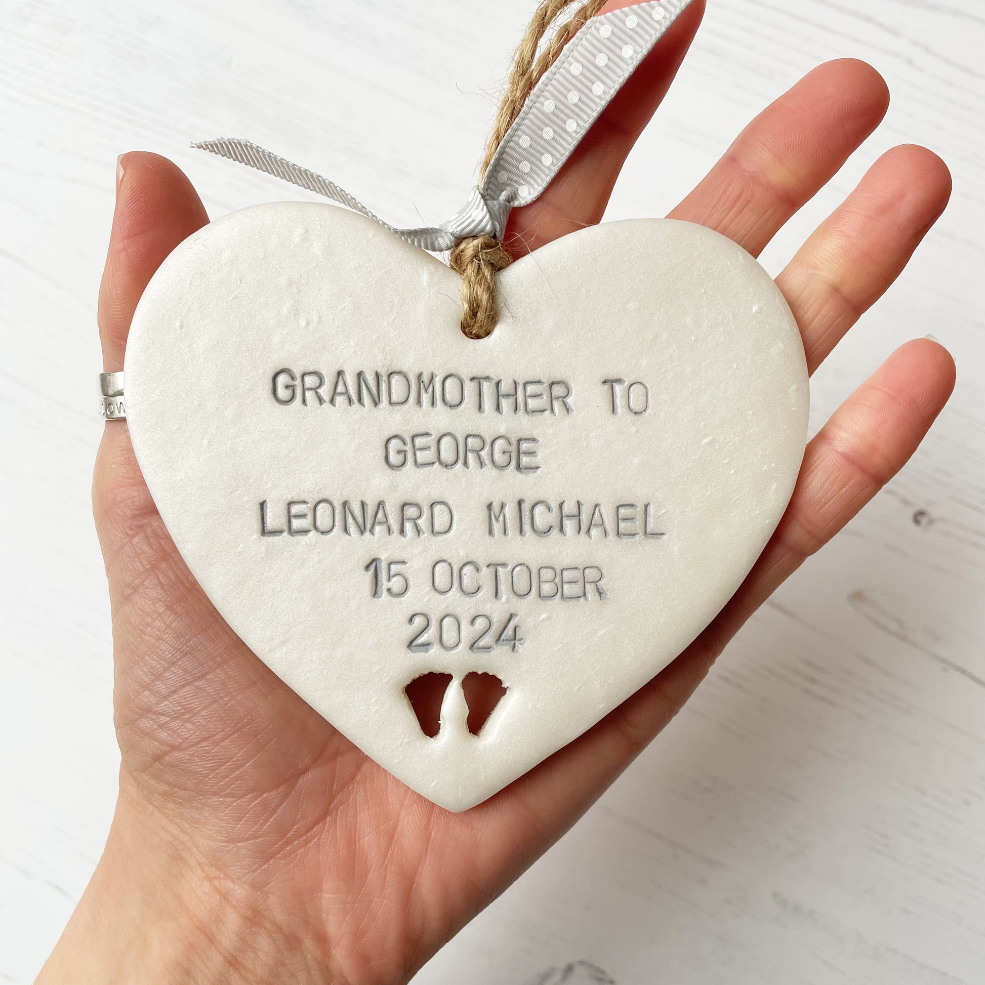 Pearlised white clay hanging heart with baby feet cut out of the bottom and grey personalisation, the heart is personalised with GRANDMOTHER TO GEORGE LEONARD MICHAEL 15TH OCTOBER 2024