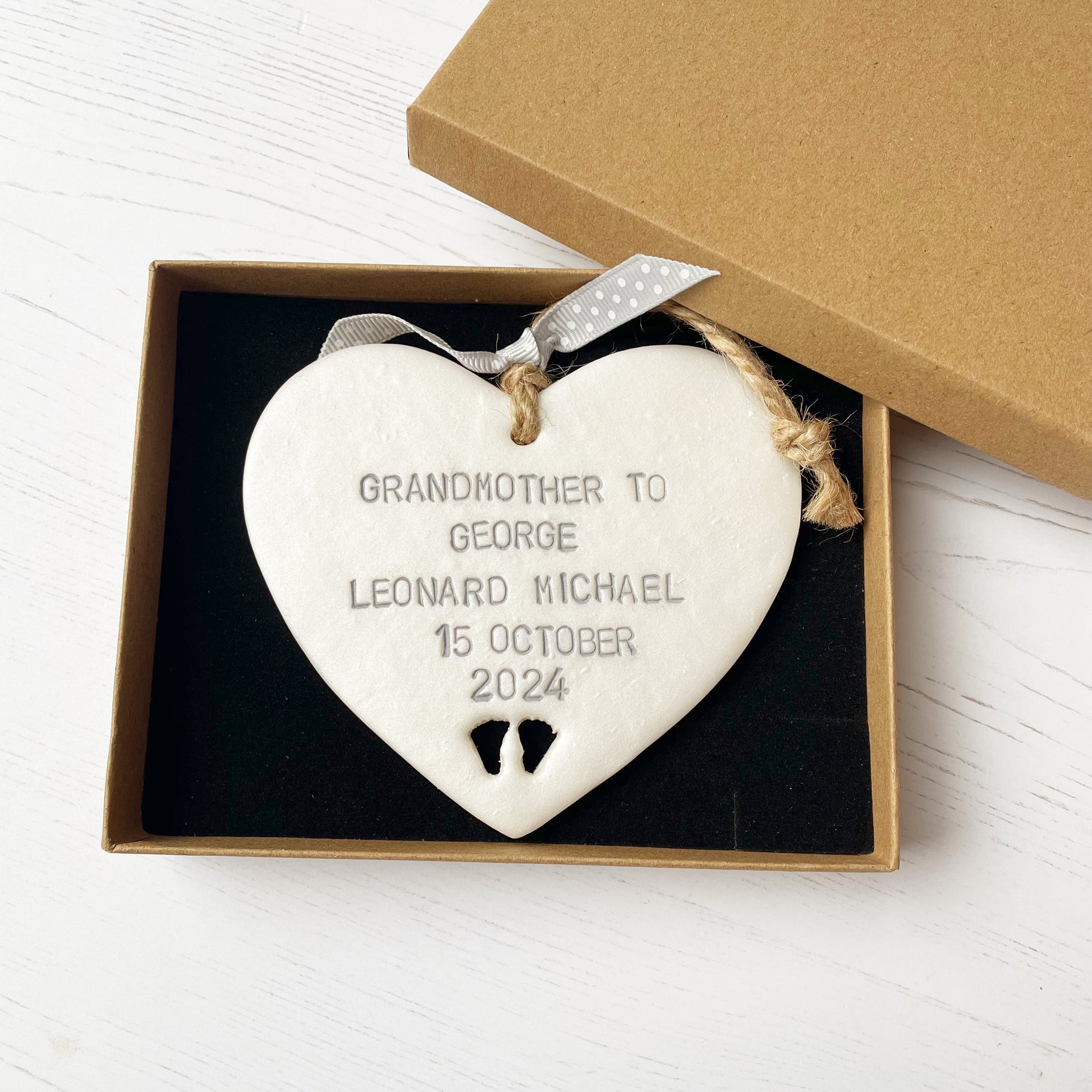 Pearlised white clay hanging heart with baby feet cut out of the bottom and grey personalisation, the heart is personalised with GRANDMOTHER TO GEORGE LEONARD MICHAEL 15TH OCTOBER 2024