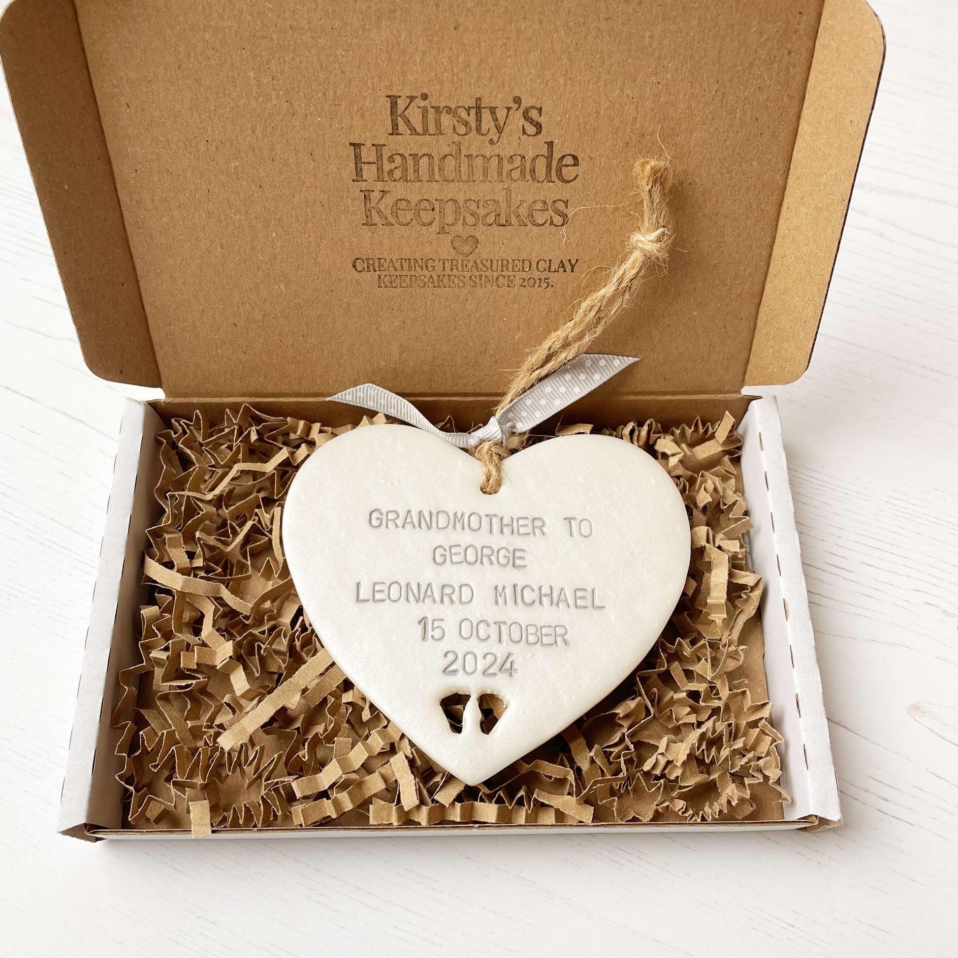 Pearlised white clay hanging heart with baby feet cut out of the bottom and grey personalisation, the heart is personalised with GRANDMOTHER TO GEORGE LEONARD MICHAEL 15TH OCTOBER 2024