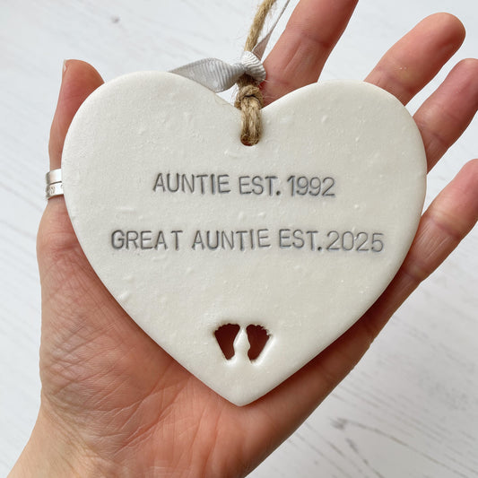 Pearlised white clay hanging heart with baby feet cut out of the bottom and grey personalisation, the heart is personalised with AUNTIE EST. 1992 GREAT AUNTIE EST. 2025