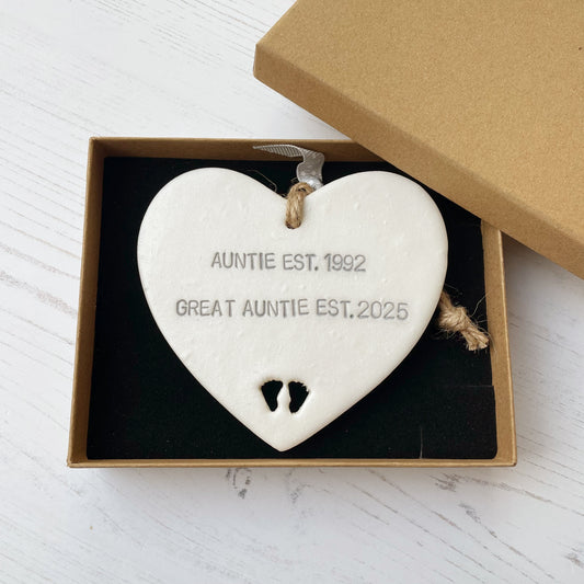 Pearlised white clay hanging heart with baby feet cut out of the bottom and grey personalisation, the heart is personalised with AUNTIE EST. 1992 GREAT AUNTIE EST. 2025