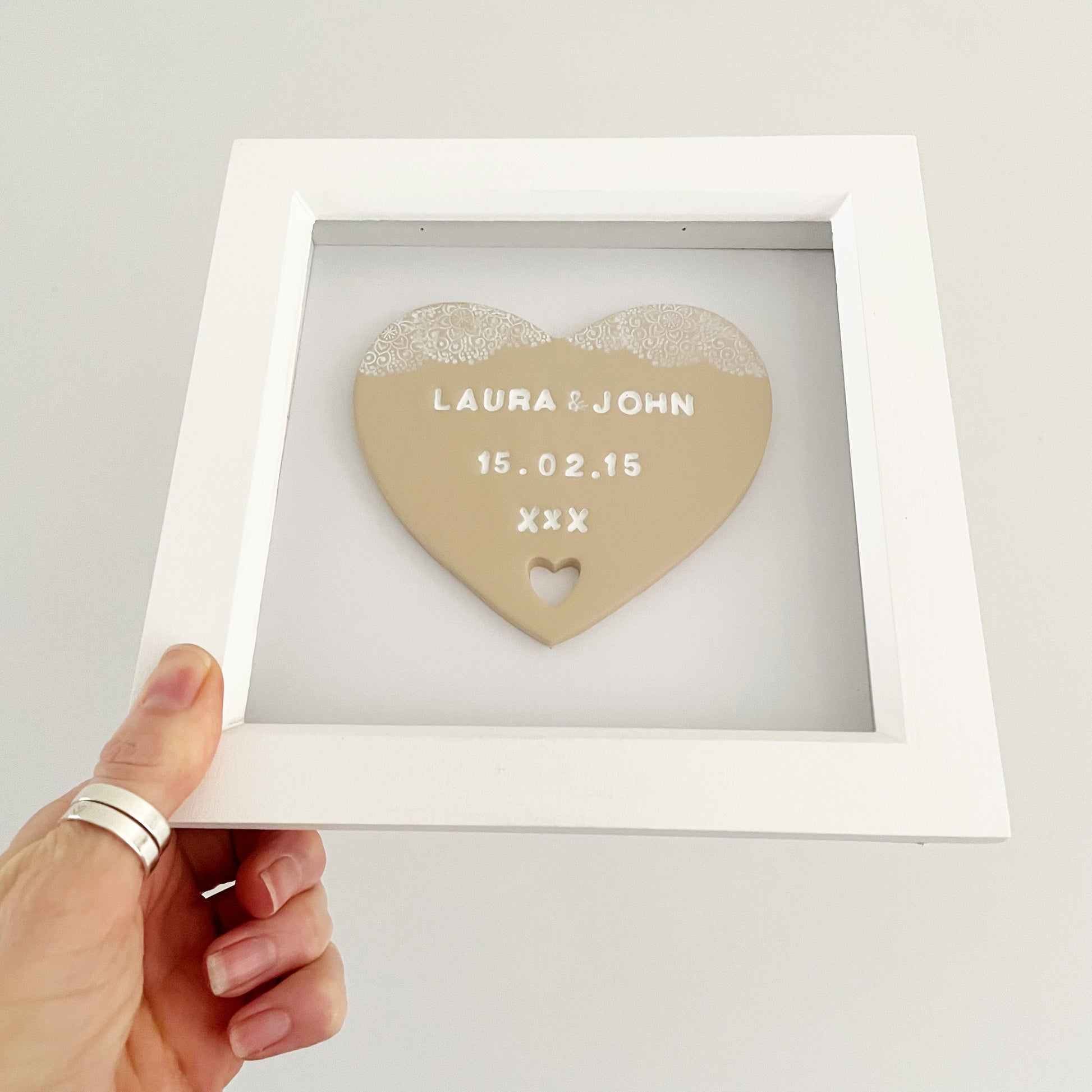 Personalised housewarming new home gift, beige clay heart with a white lace edge at the top of the heart and a heart cut out at the bottom in a white box frame, the heart is personalised with LAURA & JOHN 15.02.15 XXX