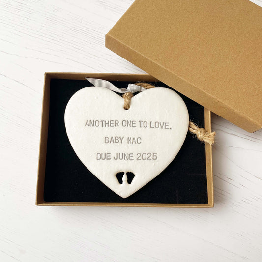 Pearlised white clay hanging heart with baby feet cut out of the bottom and grey personalisation, the heart is personalised with ANOTHER ONE TO LOVE. BABY MAC DUE JUNE 2025