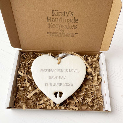 Pearlised white clay hanging heart with baby feet cut out of the bottom and grey personalisation, the heart is personalised with ANOTHER ONE TO LOVE. BABY MAC DUE JUNE 2025
