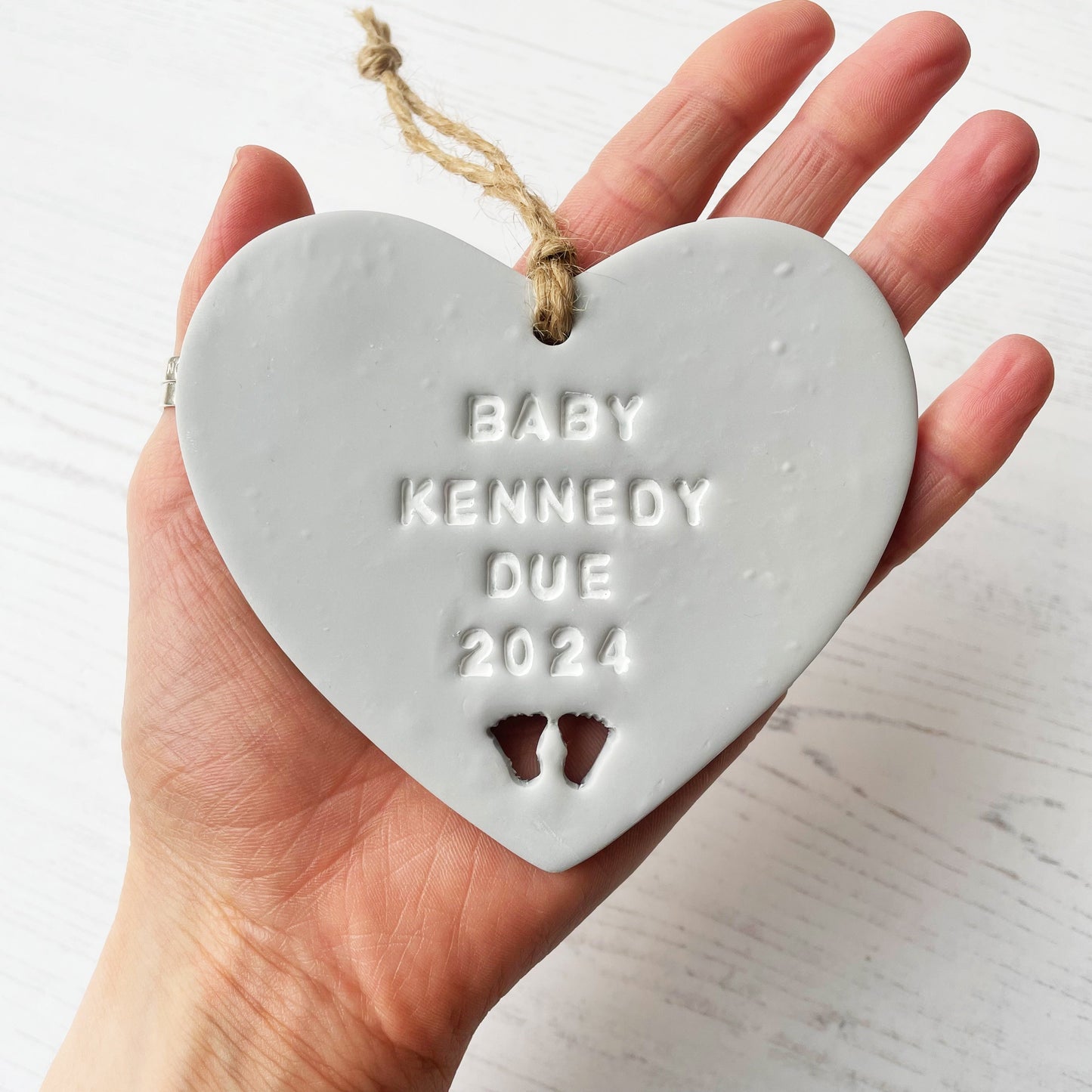 Personalised pregnancy reveal sign keepsake, grey clay hanging heart with baby feet cut out at the bottom, the heart is personalised in white with BABY KENNEDY DUE 2024