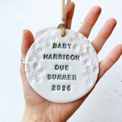 Personalised baby due Christmas bauble ornament, pearlised white round clay with BABY HARRISON DUE SUMMER 2026 in grey paint, decorated with 3 iridescent glitter snowflakes on either side of the bauble