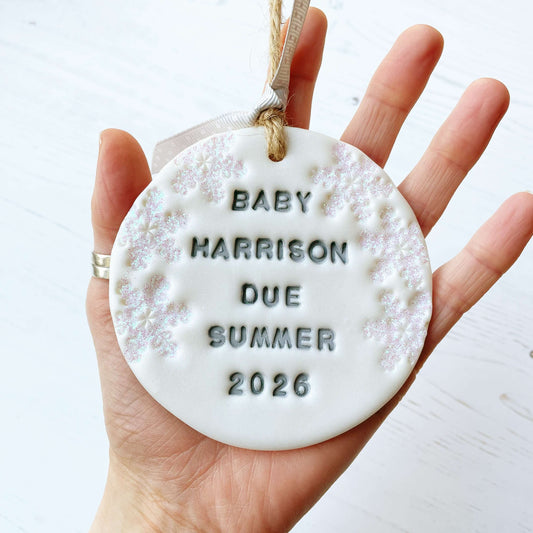 Personalised baby due Christmas bauble ornament, pearlised white round clay with BABY HARRISON DUE SUMMER 2026 in grey paint, decorated with 3 iridescent glitter snowflakes on either side of the bauble