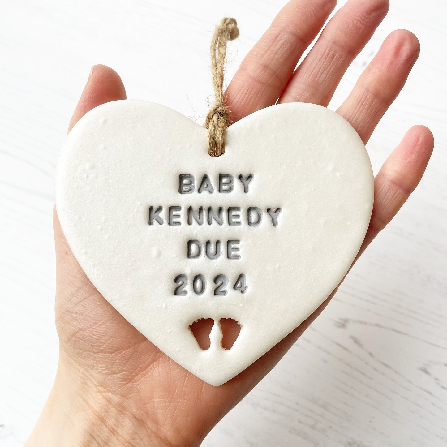 Personalised pregnancy reveal sign keepsake, pearlised white clay hanging heart with baby feet cut out at the bottom, the heart is personalised in grey with BABY KENNEDY DUE 2024