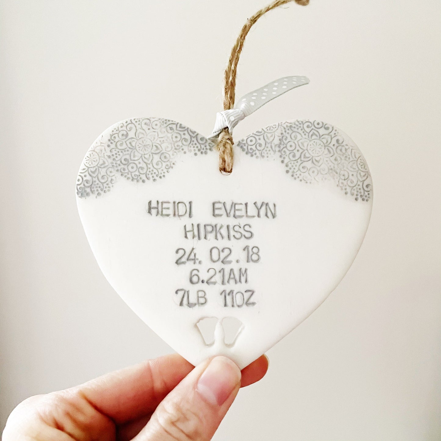 Personalised baby keepsake gift, pearlised white clay hanging heart with baby feet cut out at the bottom of the heart and a grey lace edge at the top of the heart, the heart is personalised with the baby’s name, date of birth, weight and time